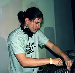 B-Complex wearing a white print t-shirt, with earmuff headphones hanging around neck and yellow reflective band on right wrist, playing a DJ control board onstage