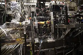 A dense laboratory setup filled with interconnected scientific instruments, cables, and metal frameworks. Optical components, valves, tubing, and control electronics surround a central experimental apparatus, with foil-wrapped sections, laser mounts, and wiring arranged across a crowded workbench in a physics or engineering research lab.