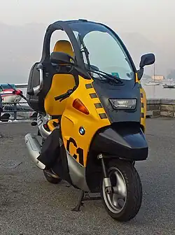 Black and yellow BMW C1 scooter parked in front of a lake with boats and an amphibian aircraft in the background