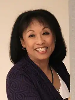 Full body portrait of a women in her late sixties smiling at the camera and with both her hands on a wall