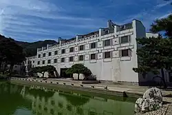 The Fragrant Hill Hotel, a white building with ornamented windows, faces a lake ringed with rock structures. Trees appear around the structure.