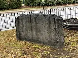 The image shows the Liberty Point Resolves Monument in Cross Creek, Fayetteville, North Carolina. The monument is a granite stone with a rounded top and jagged edges, standing on a grassy area. Centered on the smooth posterior face of the monument are the names of sixteen signers of the Liberty Point Resolves. The stone is dark gray and irregularly shaped on the sides and top. In the background, a metal fence lines a sidewalk and a road beyond it.
