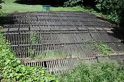 A horizontal wooden rack roughly 30 feet (9.1 m) wide and 30 feet (9.1 m) long lies in flat, grassy area.