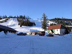 A photo of Bald Mountain Ski Area from the base of a hill. The photo shows the Lodge and Cafe to the left, rental shack, Ski-Patrol to the right. The lodge area is mid-mountain, the T-Bar base is below and to the right. The top operator's shack can be seen just right of the middle of the photo. Rope-tow and beginner's hill is between the lodge and the lower tree-line.