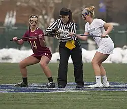 Two women, slightly hunched forward to each other, stand in the middle of an artificial turf field, while a third woman wearing a vertically black and white striped shirt stands just behind them with one hand between them