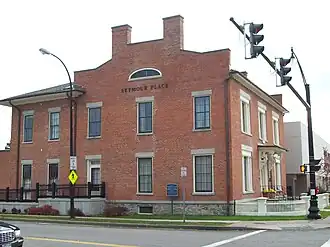 A brick building on a corner with a traffic signal in front of it. Two chimneys rise from a section that rises above the roofline on the side facing the camera; the front faces the right side. Metal lettering on the section facing the camera spells out "Seymour Place".
