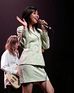Beabadoobee behind the mic, playing guitar, made-up and wearing a floral blouse.