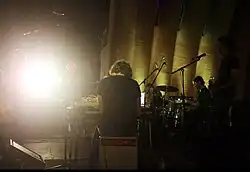 Side-stage view of Bell Gardens performing live, with instruments and microphone stands silhouetted against bright stage lighting.