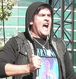 A white man with short, black hair performing on stage while holding a microphone in his right hand. He is wearing a hooded jacket over a black Nirvana t-shirt.