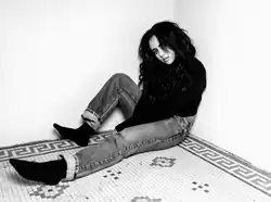 Picture of a young, brunette woman who is sat on the floor and rests on a concrete wall. She wears jeans and a dark shirt.