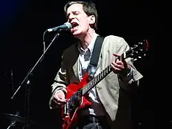 The band's frontman, performing lead vocals at the microphone, plays guitar while he sings. His face communicates strong emotion, and his posture shows great concentration on his vocal delivery.
