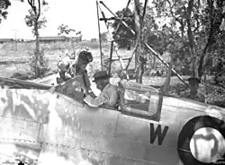 An old man wearing a hat and white shirt sitting in the cockpit of a silver aircraft. A younger man is leaning into the cockpit.