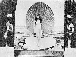 A black-and-white film still depicting a white woman in a bodysuit standing on a giant open shell against a sea background. Two white women in theatrical costumes hold the curtains.
