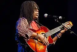 A man with hair braids is wearing sunglasses and playing a guitar.
