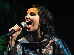 A woman wearing gold facepaint, singing into a microphone