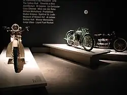 Three pristine 1920s – 1930s motorcycles on pedestals in front of text printed on the wall with words relevant to the period, such as "Prohibition" and "Al Jolson".
