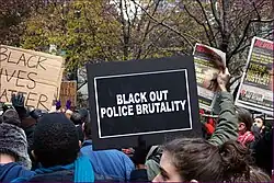 an Image of a Black Lives matter protest, including a person holding a sign which says "Black out police violence"