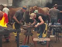 Two men lean over an anvil. One is holding down a red-hot piece of metal with tongs while the other prepares to strike with a large hammer.