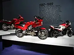 Three sporty motorcycles of the 1990s on pedestals. Text on the wall behind them says "1982–1989" with a few dozen words relevant to the period, such as "Atari", "MTV", and "Robert Mapplethorpe".