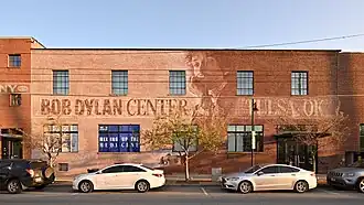 Exterior view of the Bob Dylan Center in Tulsa, Oklahoma. Painted mural depicting folk musician Bob Dylan between text that says Bob Dylan Center and Tulsa, OK, on a brick wall.