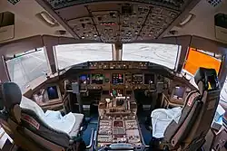 A flight deck, from behind the two pilots' seats. A center console lies in between the seats, in front is an instrument panel with several displays, and light enters through the forward windows.