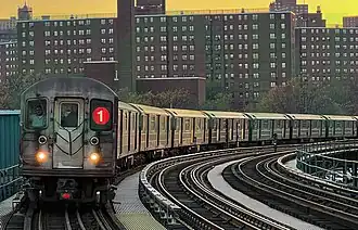 A "1" train, composed of R62A cars, is seen above ground entering the 207th Street station. The front of the train contains two white lights providing slight illumination, two windows, a door, and the Symbol for the 1 line on the left window.