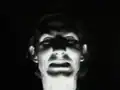 Black-and-white video still of a performer's face lit from below, creating a dramatic contrast.