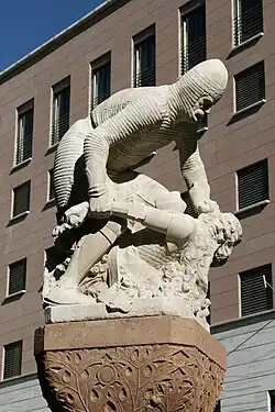 Photograph of a sculpture in white stone. A leaning man in chain mail pins down a smaller bearded man. Below the sculpture, there is a red stone carved capitel. In the background, a modern building with more than four storeys.