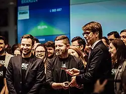 A group of about a dozen elegantly dressed young people stand on stage during a stock exchange event; two of the three men in the foreground are passing something to each other, while in the background you can see a screen showing the company's rising stock prices.