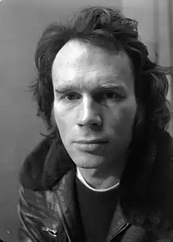 A black-and-white portrait of a man with wavy hair wearing a leather jacket with a fur collar, gazing directly at the camera with a calm, serious expression.