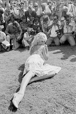 A black and white photograph of a woman posing while laying on the ground. In front of her, numerous photographers take pictures of her.