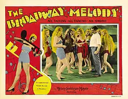 A lobby card with a man surrounded by smiling showgirls in long wigs, framed by a red musical border with a dancer and illustrated musical notes.
