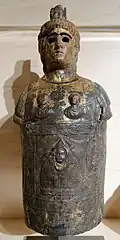 Photograph of an ancient bronze statue with a sculpted face, intricate details, and depicted figures. The surface is weathered, evoking a sense of historical significance.