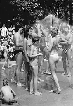 A black and white photograph from Berlin in 1987, showing a group of people using an outdoor shower. The photo shows two shirtless men, two topless women and two children.
