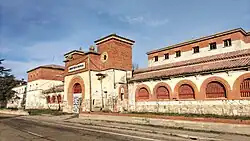 Photograph of a two-storey building with white walls. The gate and the windows are topped by brick arches. The windows have been bricked up. There is graffiti on the walls.