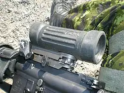 Close-up of a C79 optical sight viewed from the left side, showing the rectangular prism housing, textured adjustment knobs, and mounting base attached to a rifle rail.