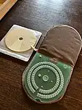 green plastic slate for adding braille to a CD sits inside of a brown fabric cover. Next to the slate are circular pieces of paper the size of a CD.