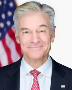 A shoulder up portrait of Mehmet Oz. He is smiling and looking directly into the camera. He is wearing a dark navy suit, a white shirt, and a red tie with a blue floral and horseshoe print.