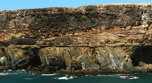 Cliff at Caleta Negra, Ajuy on Fuerteventura. The lower third of the cliff consists of steeply-tilted pre-Miocene oceanic sedimentary rocks. These rocks are unconformably overlain by Pliocene light-coloured cross-bedded beach sedimentary rocks, black pillow lava, and a black lava flow.[310][311] (Scale: cliff height is approximately 40&nbsp;m (130&nbsp;ft)[312])