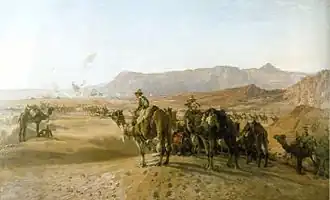 A large number of men mounted on camels in a treeless desert. There is a town in the distance, and bare rocky mountains beyond.