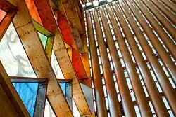 Close-up architectural details showing cardboard tubes, wooden elements, and colored glass panels in clear, red, yellow, green, and blue