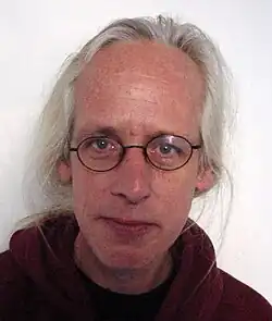 Man with long gray hair, round glasses, and a maroon hoodie looking directly at the camera against a plain background