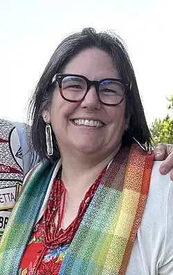 This is a photo of Carmen Lansdowne. She is a middle aged indigenous women with dark hair speckled with grey hair. She is wearing a rainbow clergy stole.