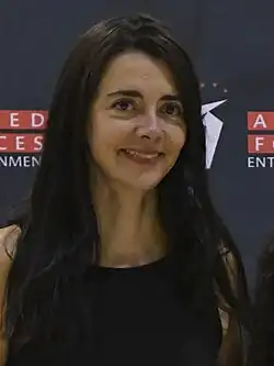 A woman with long dark hair and a black sleeveless top smiles in front of a dark backdrop with red and white text