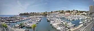 A wide panoramic view of Carry-le-Rouet harbour in France, showing a marina filled with boats, palm trees lining the waterfront, and surrounding coastal buildings under a bright sky.
