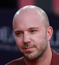 A close-up of a handsome bald man with light stubble and a calm, thoughtful expression, wearing a maroon jacket