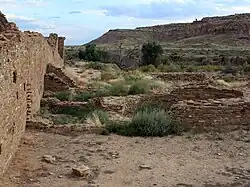 A partly overcast sky and subdued sunlight over a roughly six-foot-tall wall of dusky tan sandstone bricks which vary somewhat in size. The wall runs diagonally from the immediate foreground at left towards the right, running perhaps several dozen feet to the near middle distance. A few feet to the right, in the middle foreground, a low ring of similar blocks delimits a circular pit sunk into the ground. The remains of several other ruinous low walls, perhaps one to three high at most, are arrayed in parallel; they align left to right from the high diagonal wall. Perhaps a mile distant to the center and right, a canyon wall slopes gradually level to meet the valley floor on which the walls sit.