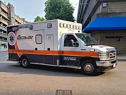 A white ambulance with orange and blue markings drives on a city road.