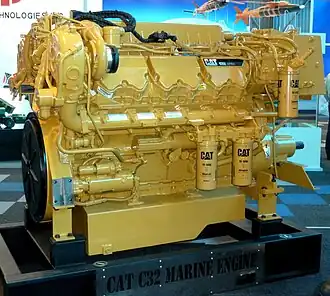 Caterpillar engine. On the right, there is a cylindrical oil filter and on slightly below the middle left also two oil filters side by side. "CAT 1R-1808 - Advanced" reads on these filters. On the bottom, it reads "CAT C32 MARINE ENGINE". On the upper right there is a black plate reading "CAT C32" along with some unreadable text. Several white quality check stickers are glued on the engine.