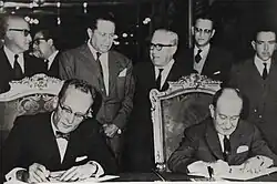 A black and white photograph of two mature and formally dressed men seated in chairs, signing some papers over a large table while a group of six men in suits stand behind their seats.